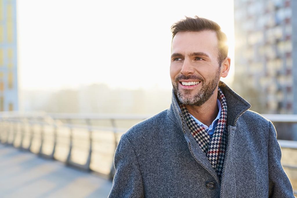 smiling man with houndstooth scarf and tweed jacket stands on walkway on his way to medical detox program