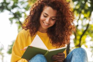 happy curly hair woman in yellow sweater writing in journal under trees during Cognitive Behavioral Therapy Program